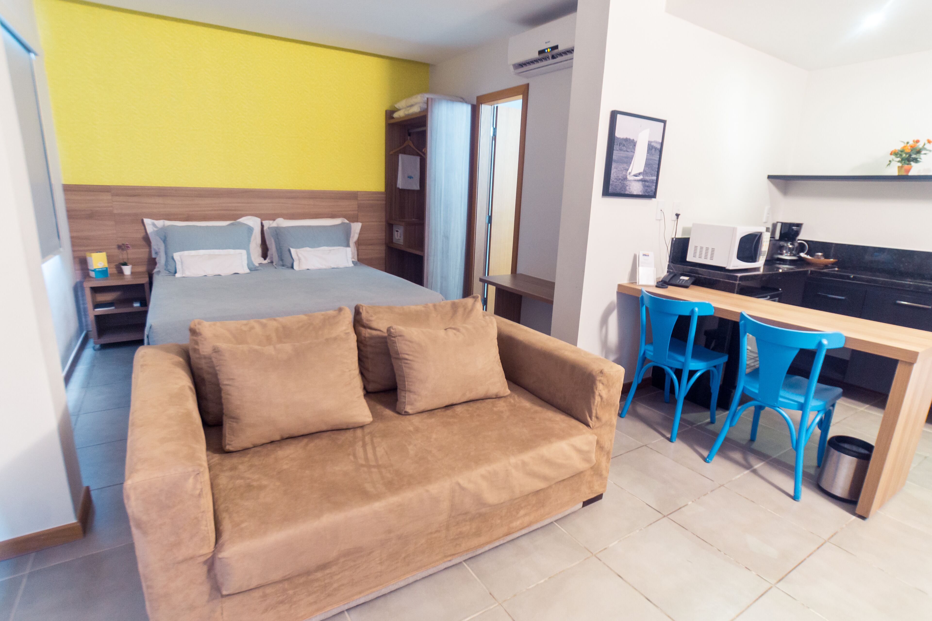 A bedroom with a bed, a sofa, a desk with chairs, and a kitchenette.