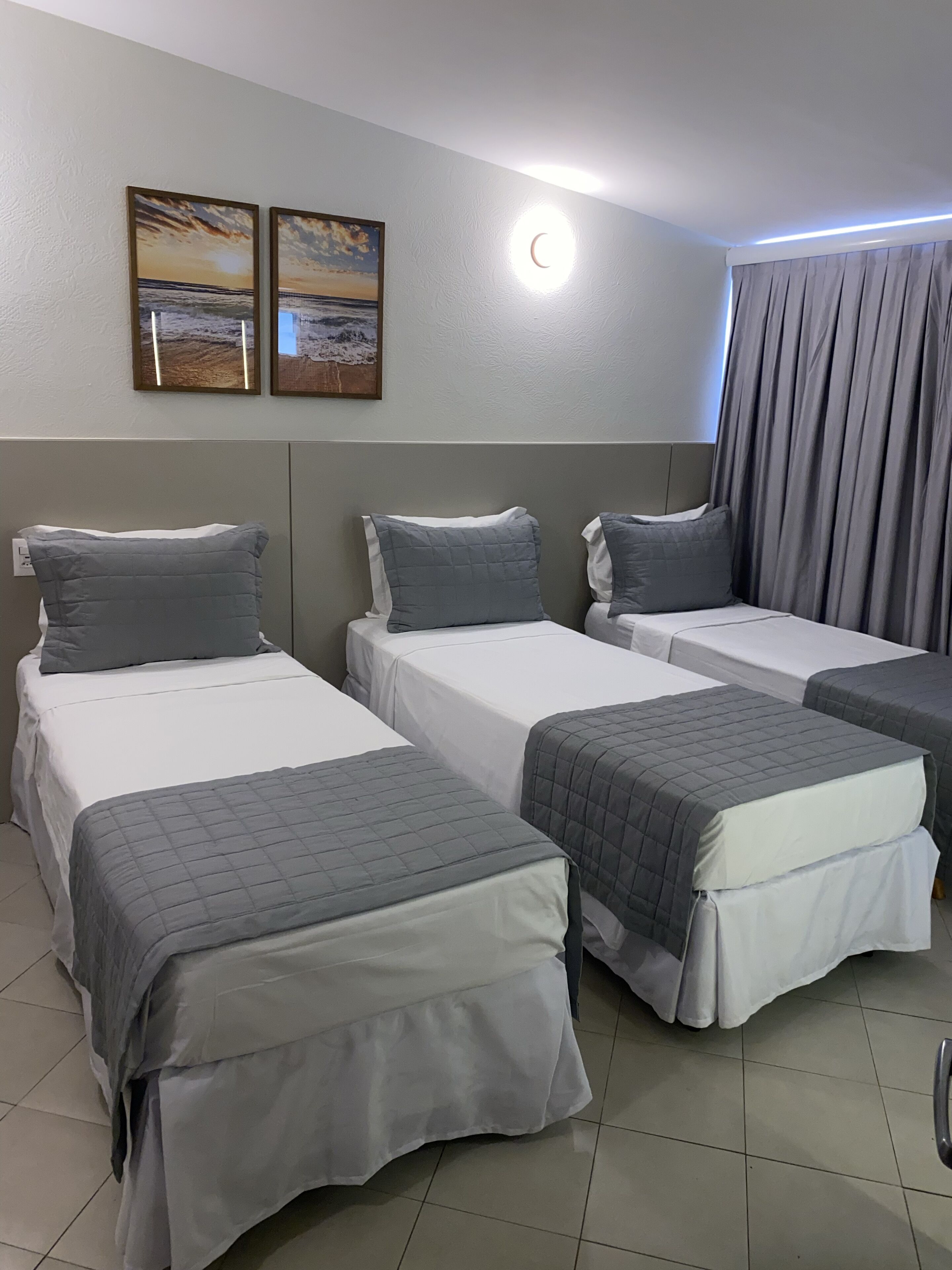 A hotel room with three beds, each with a grey quilted bedspread and pillows. There are two framed pictures on the wall above the beds.