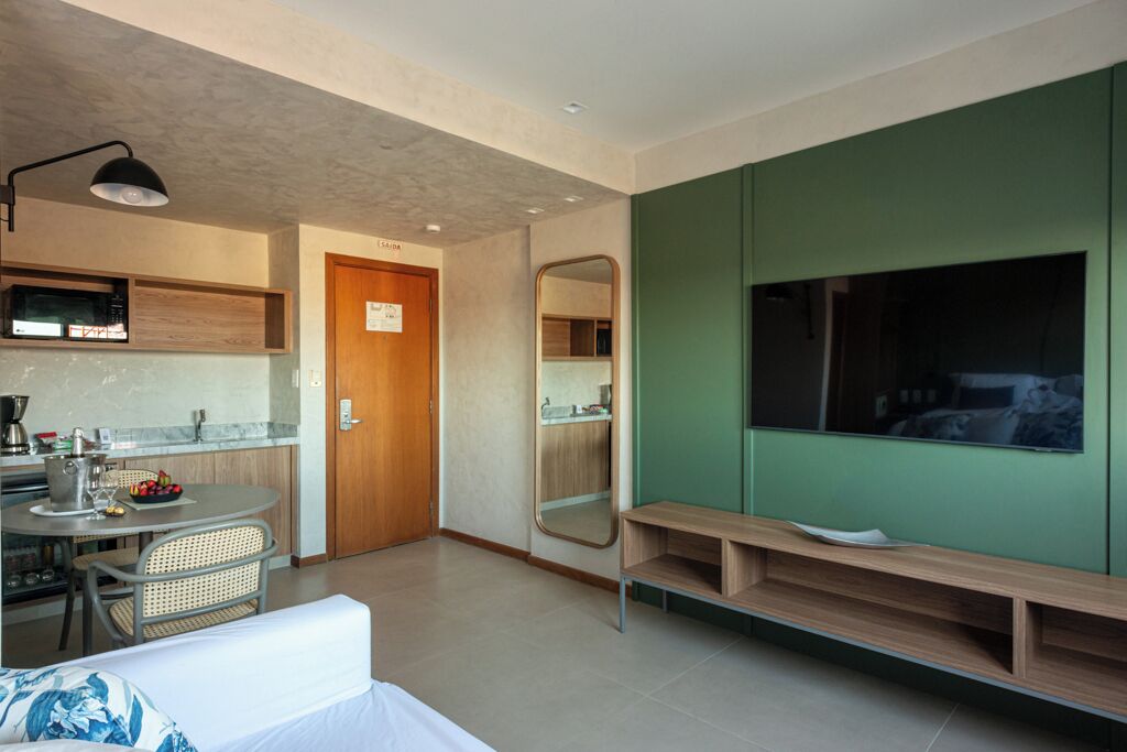 Uma sala de estar moderna com uma parede de destaque verde, um espelho grande, uma TV, um banco de madeira e uma área de jantar com mesa e cadeiras.