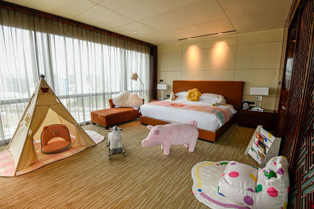 Quarto de hotel com uma cama grande, uma tenda, um brinquedo em forma de porco e uma cadeira com almofadas coloridas.