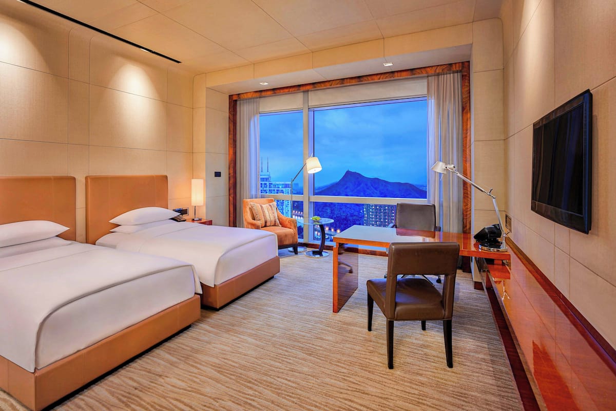 A hotel room with two beds, a desk, a chair, a TV, and a large window with a city view.