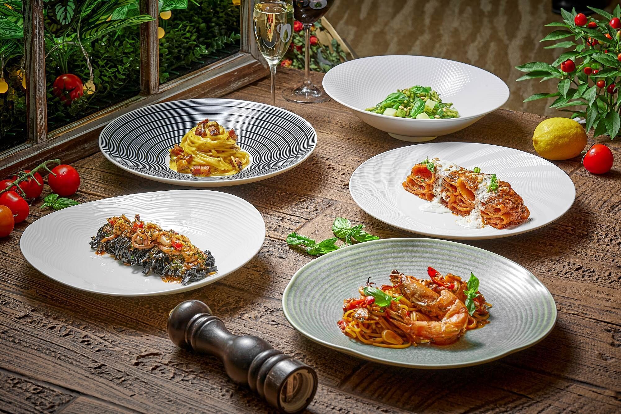 A wooden table with five plates of pasta dishes, a pepper shaker, and a glass of wine.