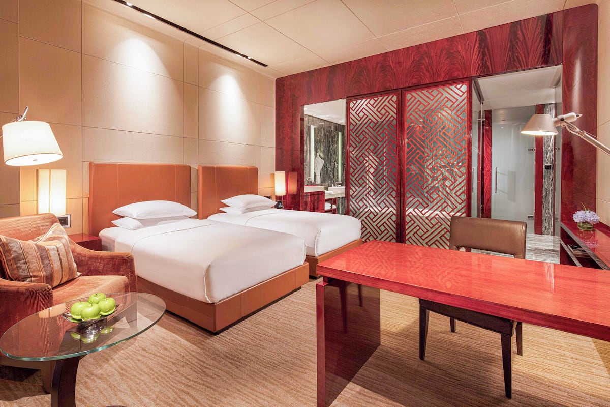 A hotel room with a bed, a glass table with apples, a red table, a brown leather chair, and a mirror.