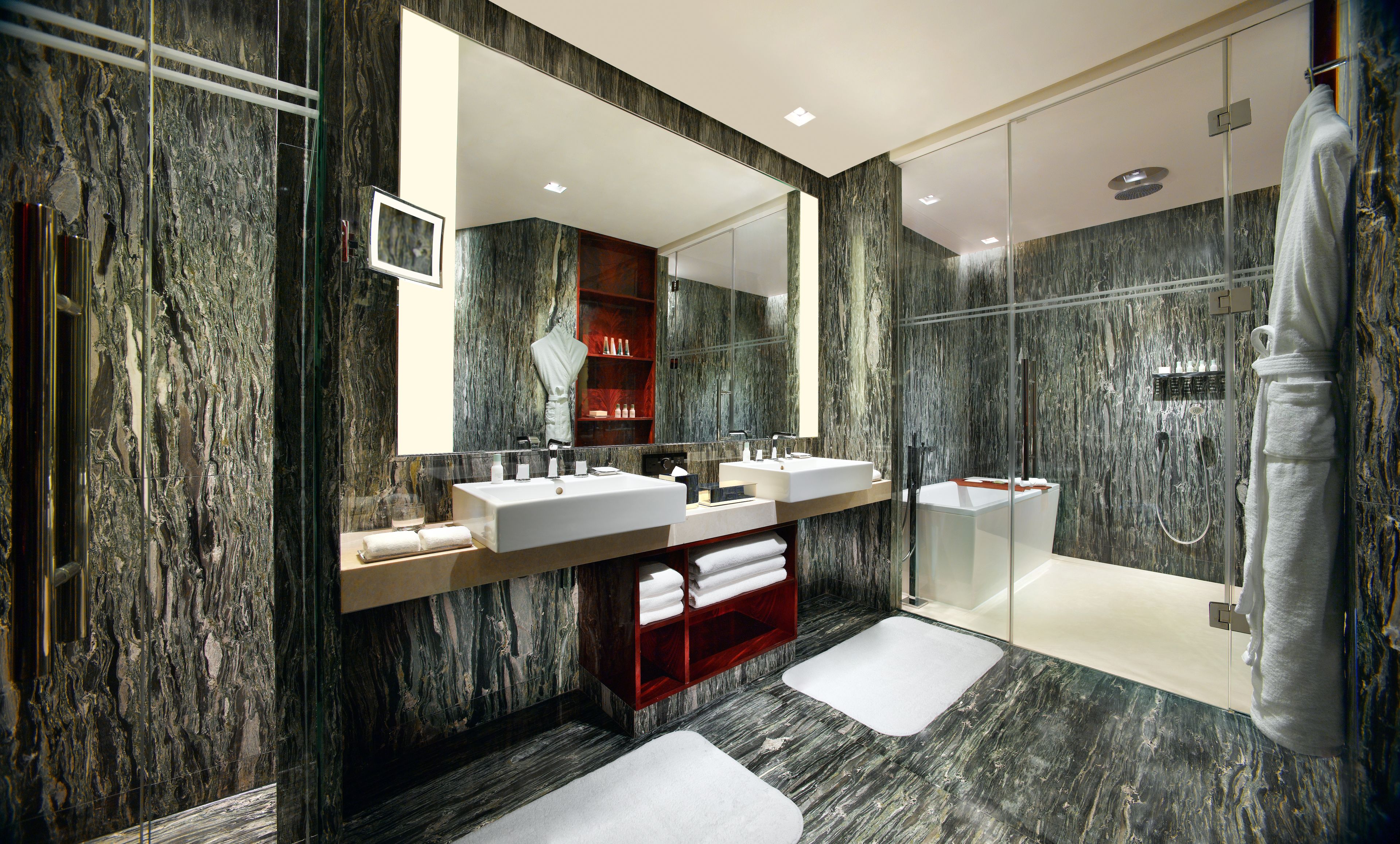 A modern bathroom with marble walls, dual sinks, a glass shower enclosure, and a red vanity.
