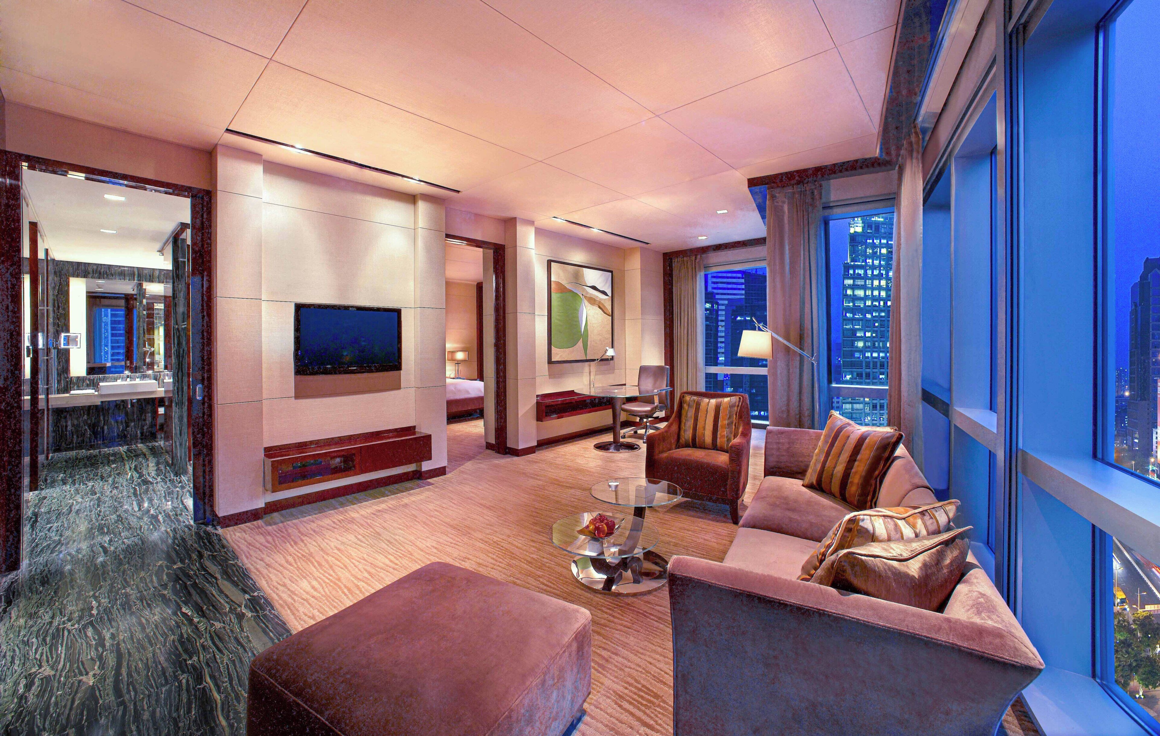A modern hotel room with a large TV, comfortable seating, a glass coffee table, and a view of the cityscape.