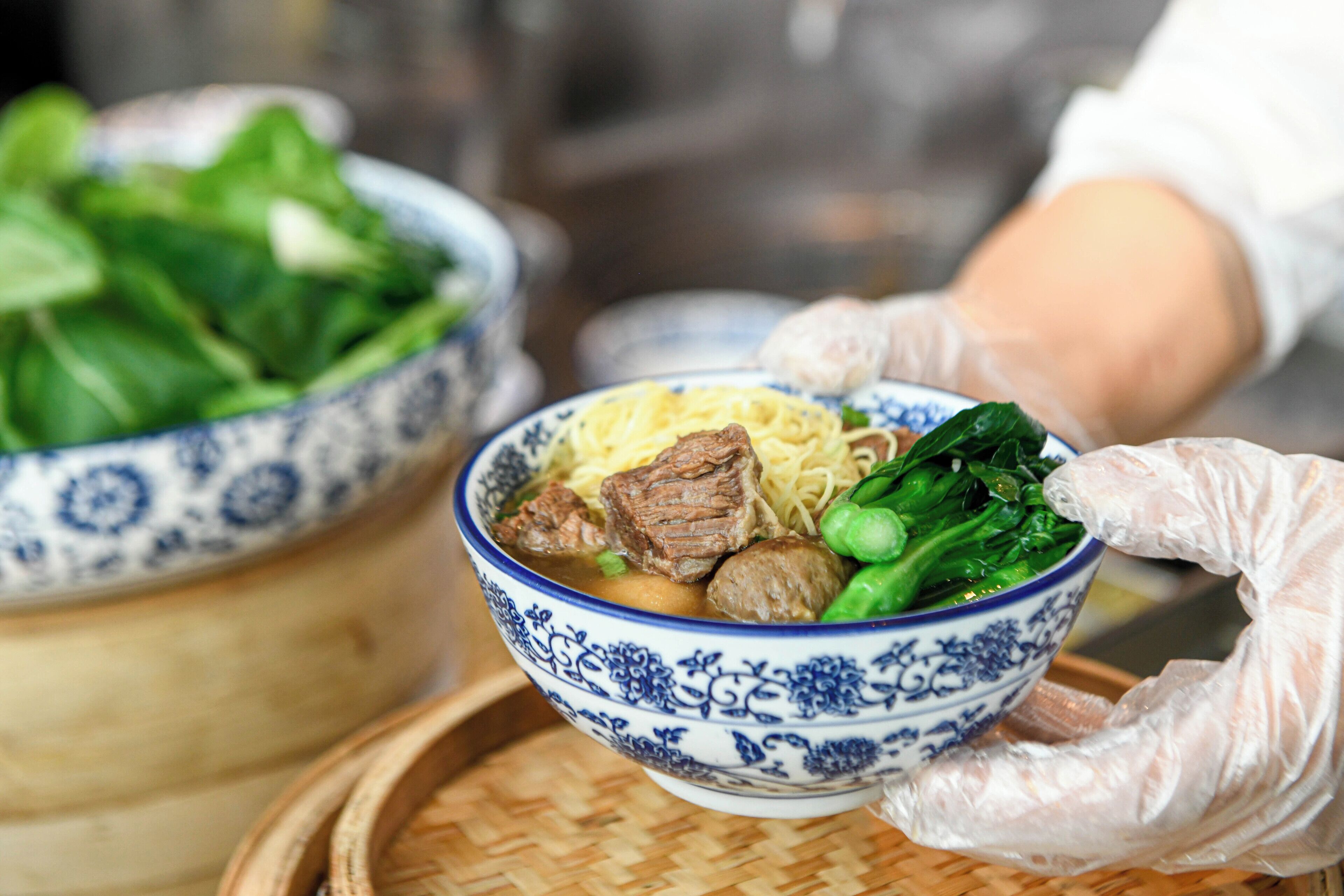 A bowl of noodle soup with beef and greens, held by a gloved hand.