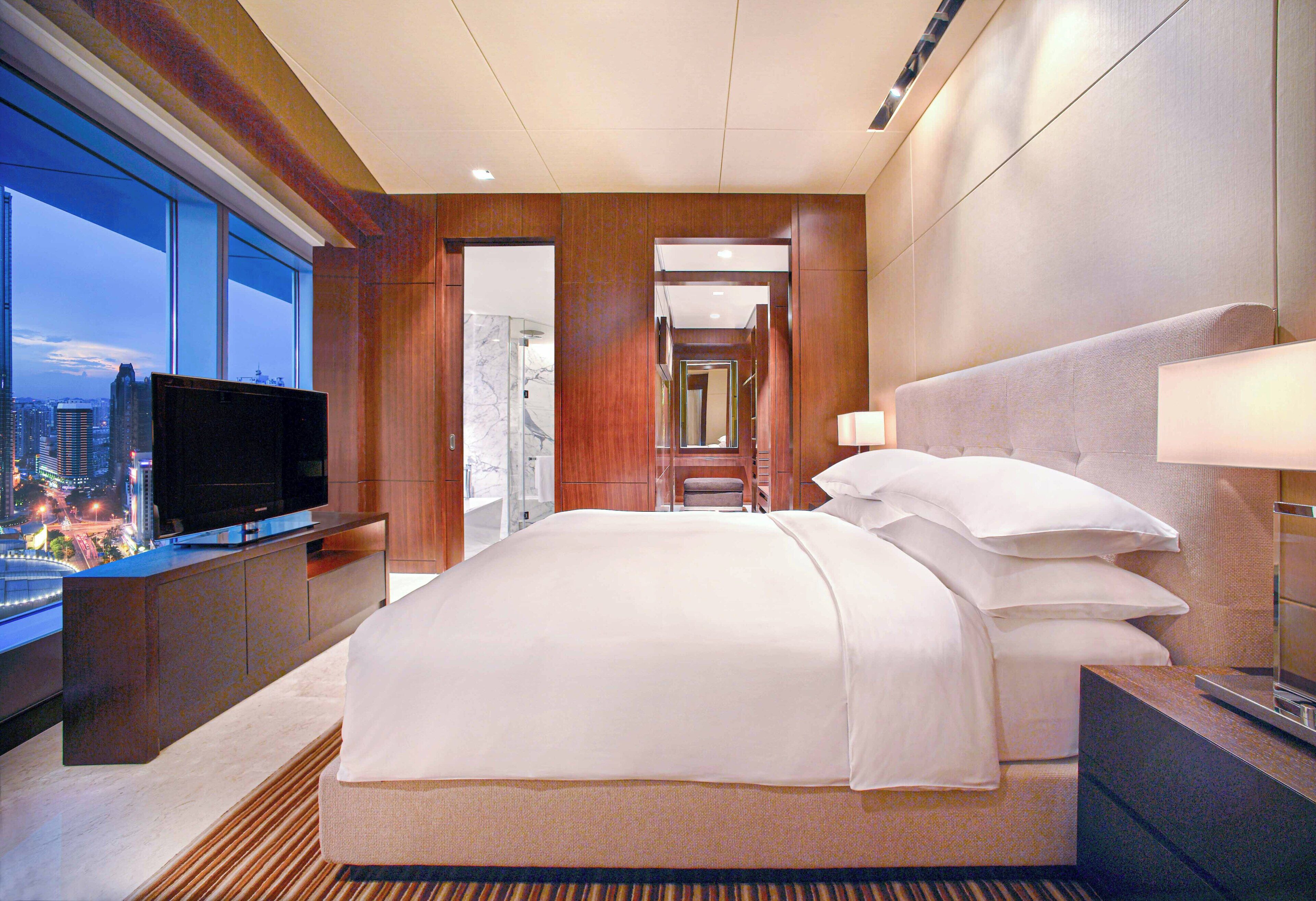 A hotel room with a large bed, a television on a stand, a nightstand, and a view of the cityscape through large windows.
