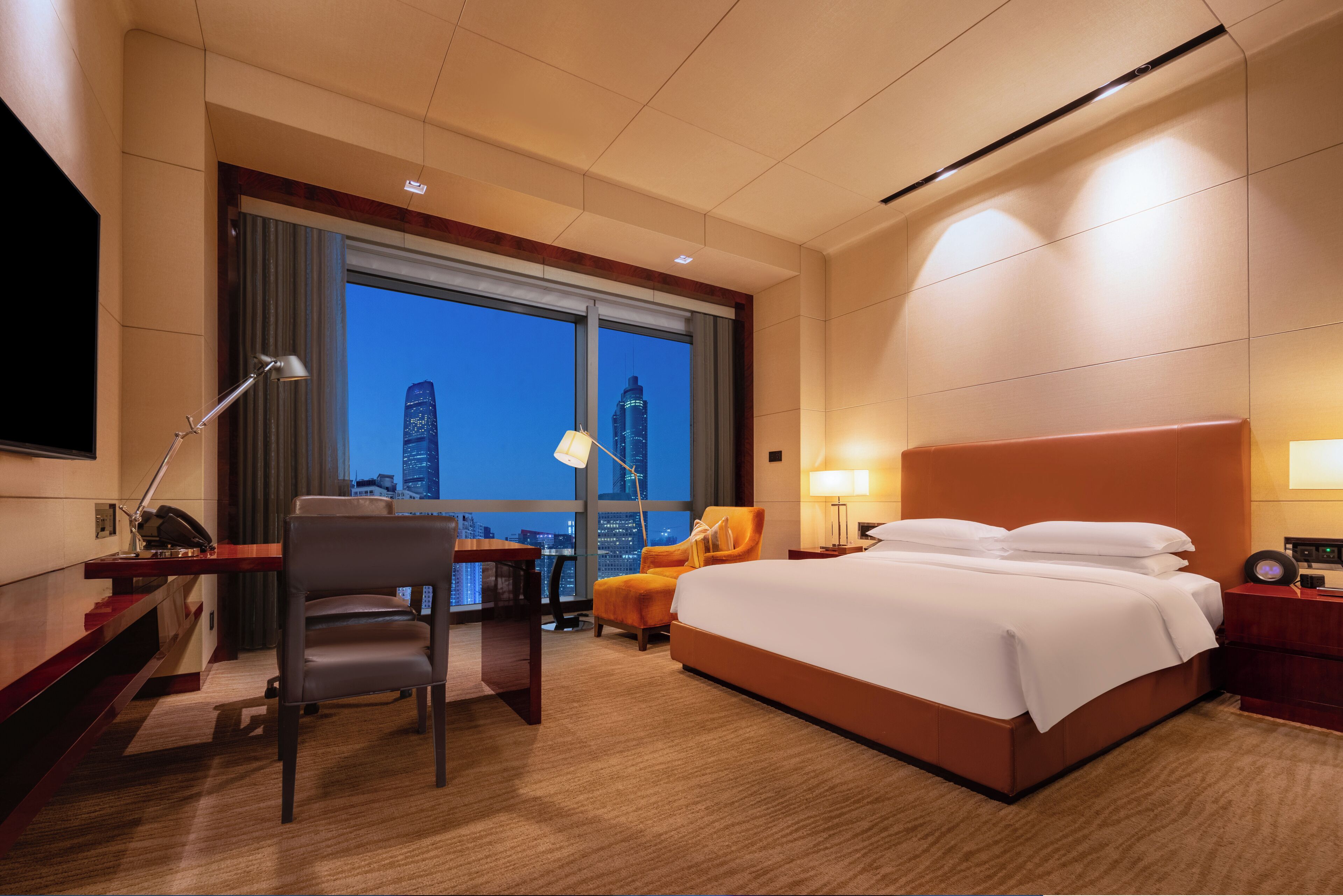 A hotel room with a large bed, a desk with a chair, a desk lamp, a nightstand, and a view of the city skyline at dusk.