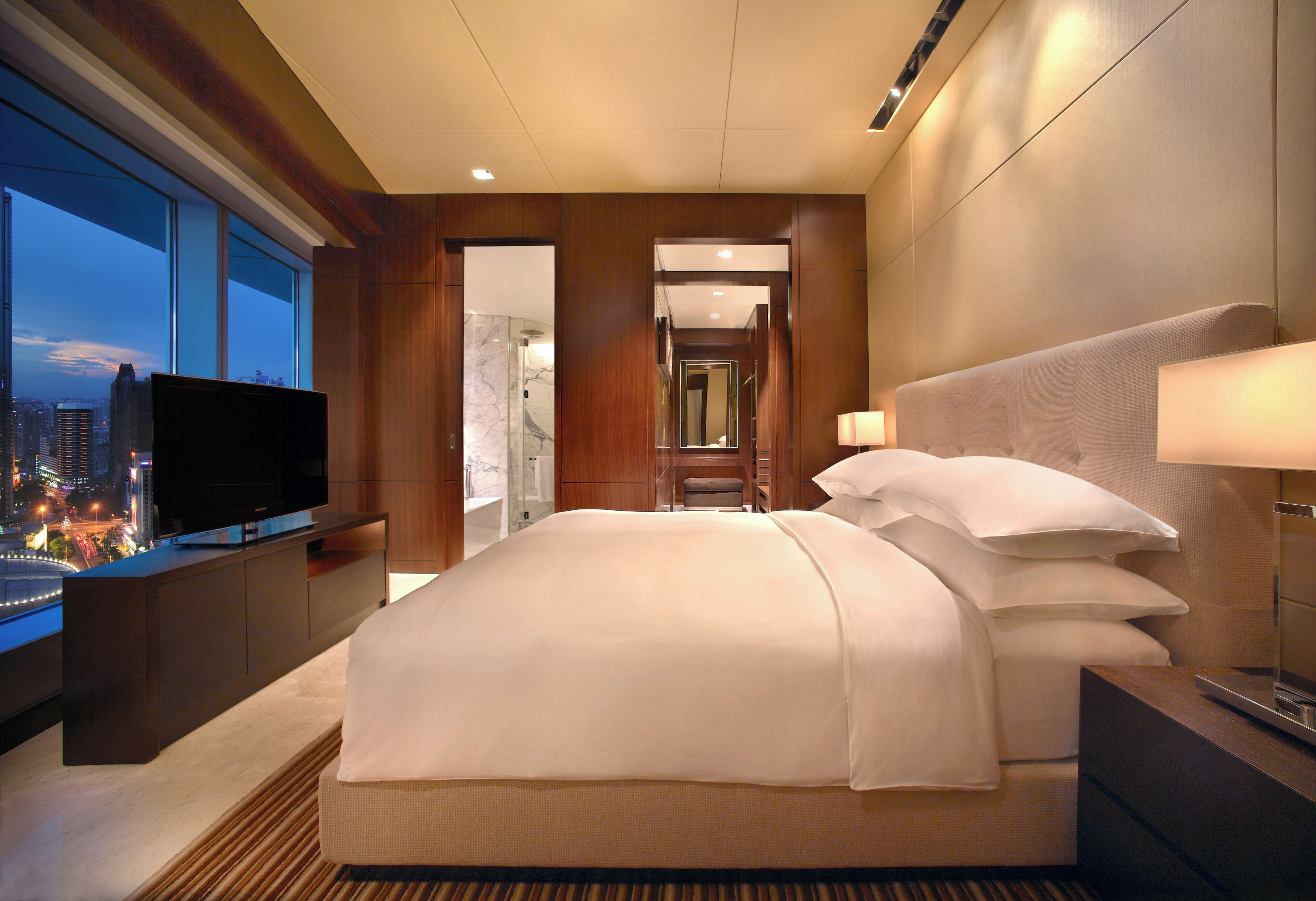A modern hotel room with a large bed, a television, and a view of the cityscape.