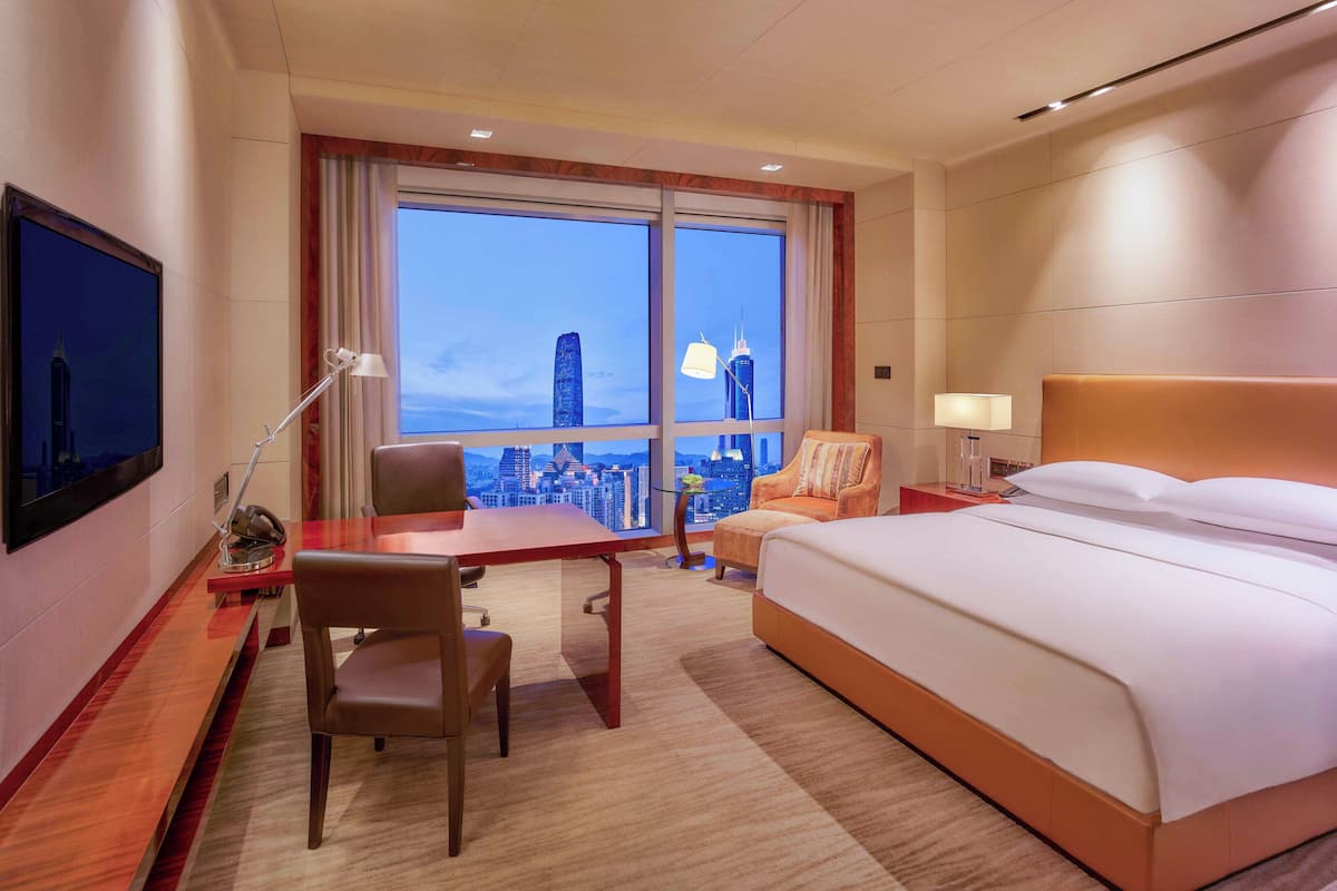 A hotel room with a large bed, a desk, a chair, a TV, and a view of a cityscape through large windows.