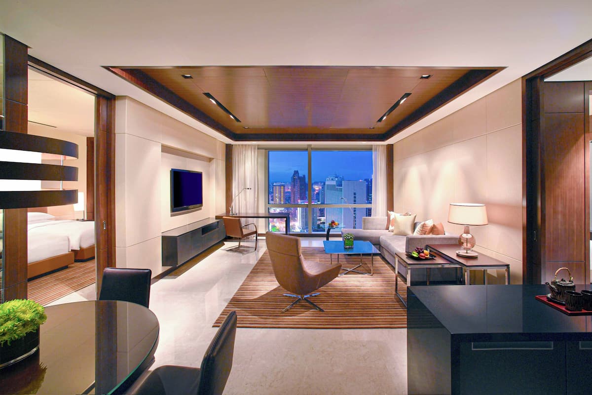 A modern hotel room with a city view, a flat-screen TV, a sofa, a dining table, and a small balcony.