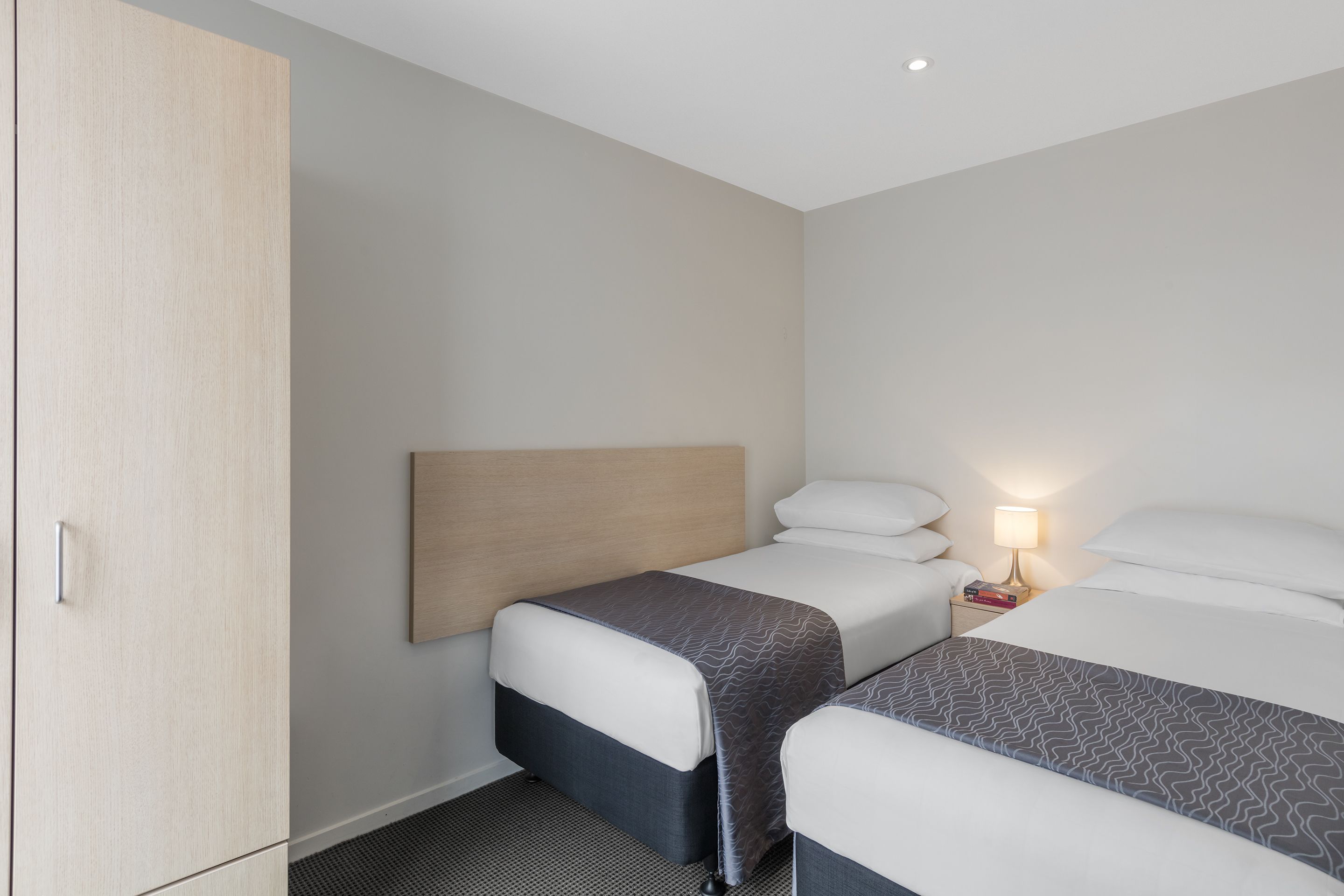 Une chambre d’hôtel avec deux lits, une armoire, une table de chevet avec une lampe et un sol recouvert de moquette.