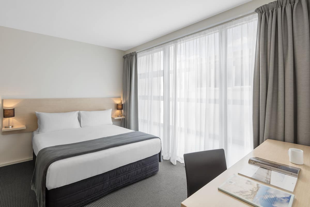 A hotel room with a bed, bedside lamps, a desk with a chair, and a large window with curtains.
