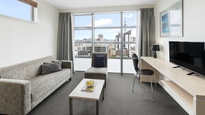A modern hotel room with a sofa, a small table with a bowl of fruit, a TV, a desk, and large windows offering a city view.