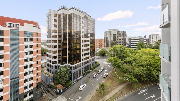 A modern high-rise building with a glass facade, surrounded by other buildings and a tree-lined street.