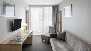 A hotel room with a flat-screen TV, a desk, a sofa, and a window with sheer curtains.