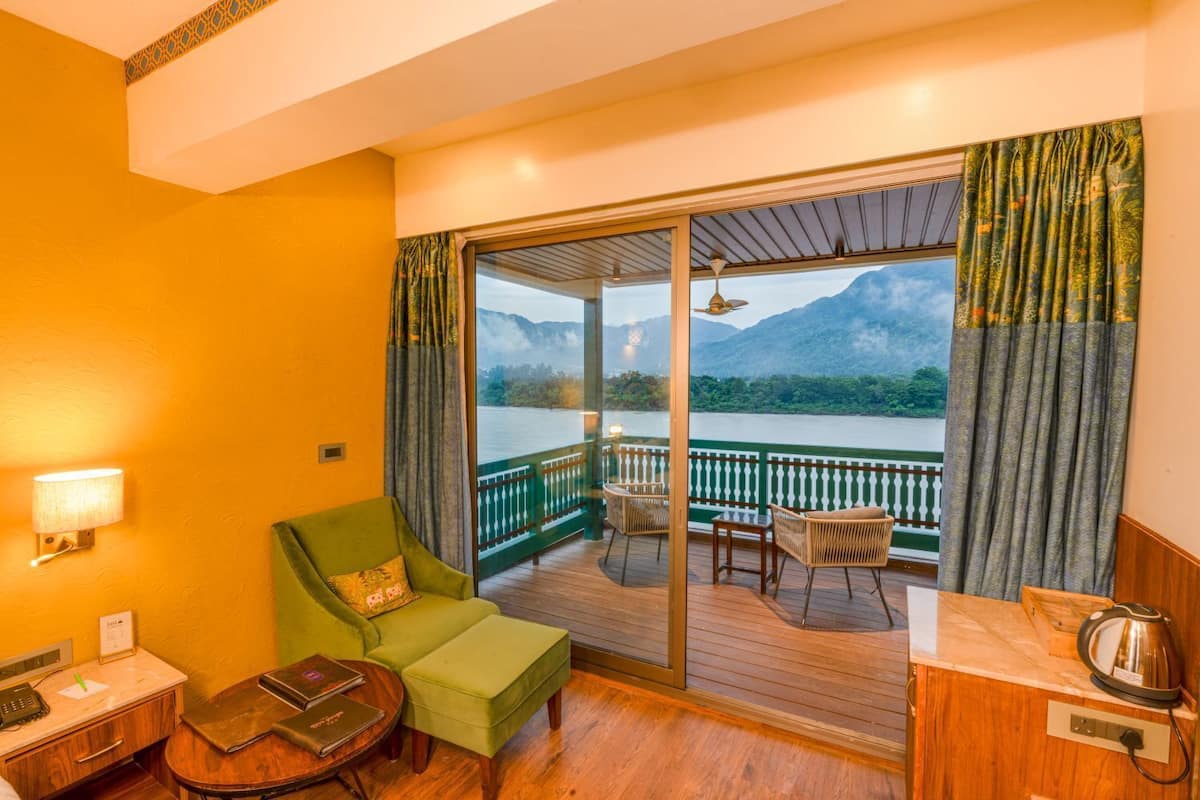 Una habitación con un sillón verde, una mesita pequeña, un escritorio de madera y vistas a un lago con montañas al fondo.