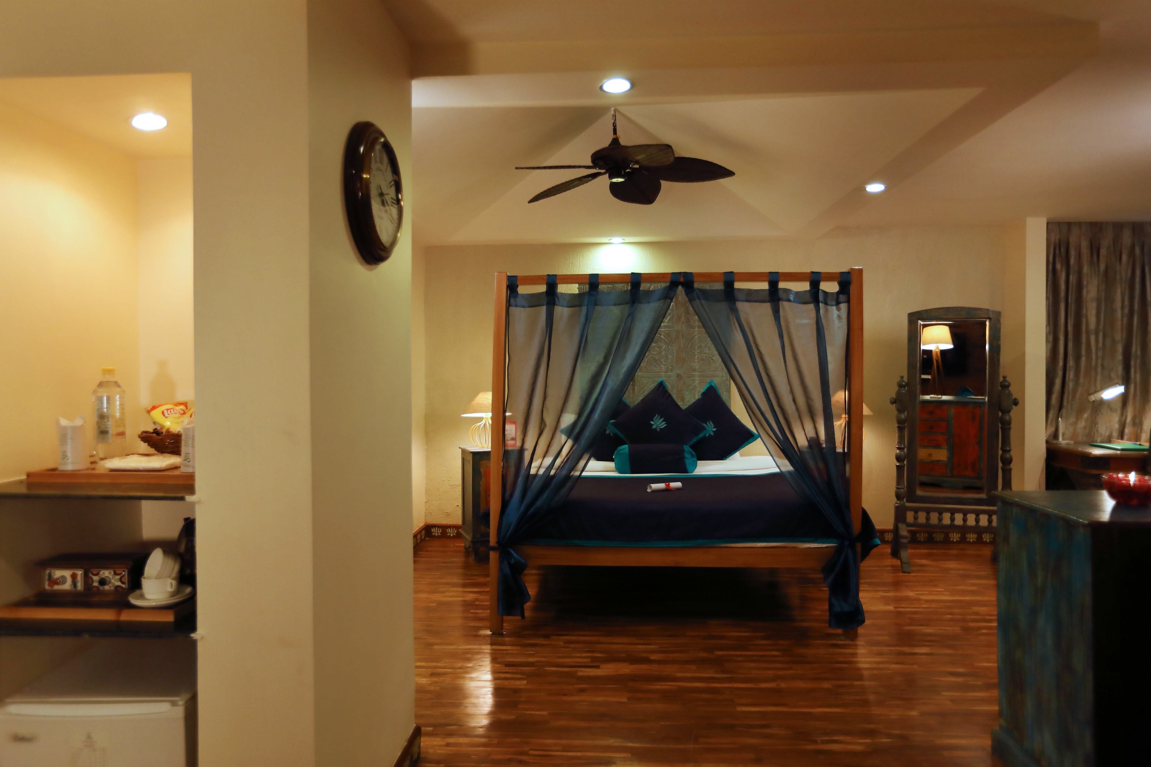 A bedroom with a bed, a ceiling fan, a clock, a nightstand, a lamp, and a wooden floor.
