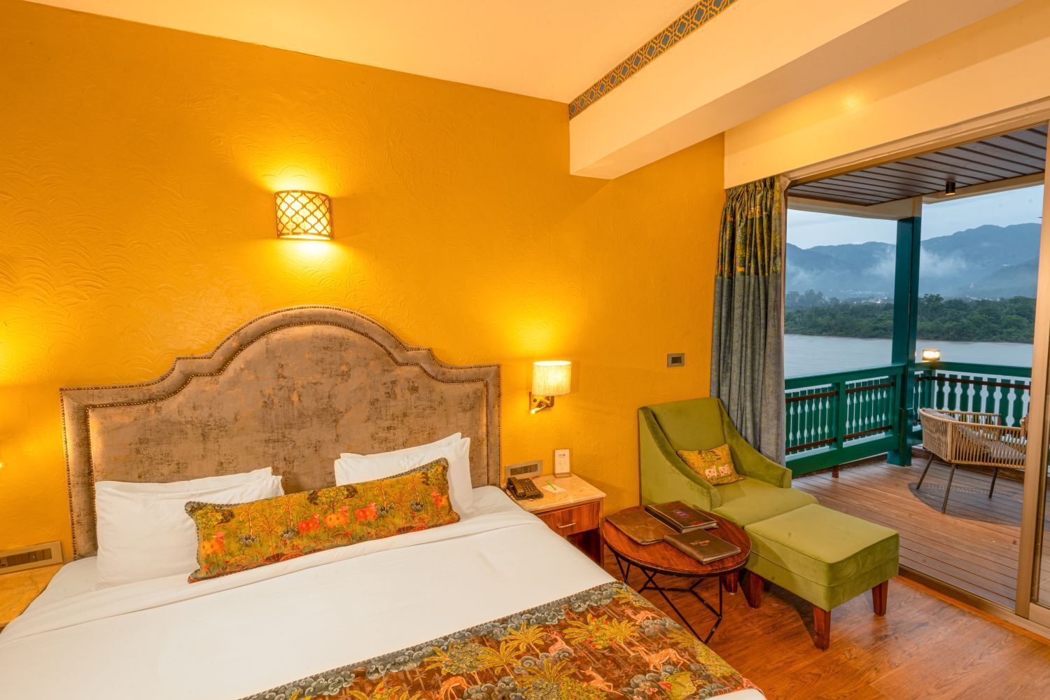 Une chambre d’hôtel avec un grand lit, un fauteuil vert, une petite table et un balcon offrant une vue sur les montagnes.