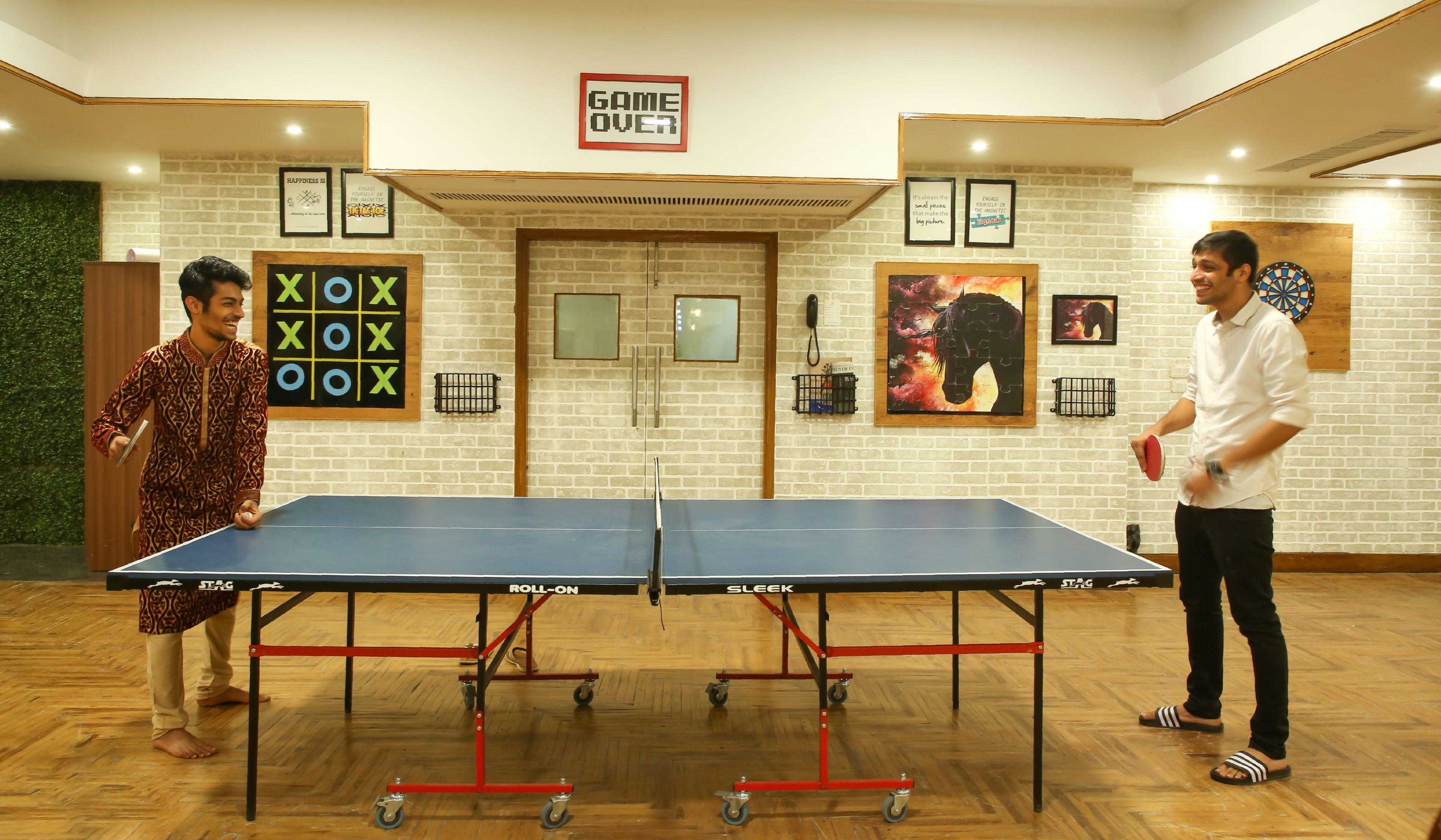 A ping pong table in a room with a brick wall, a dartboard, and motivational posters.