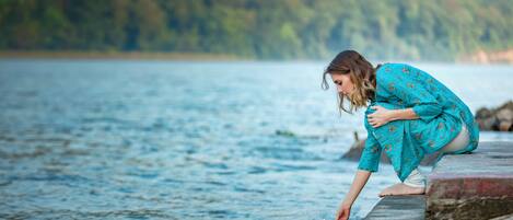 A person in a blue patterned dress by a body of water, kneeling on a concrete ledge.