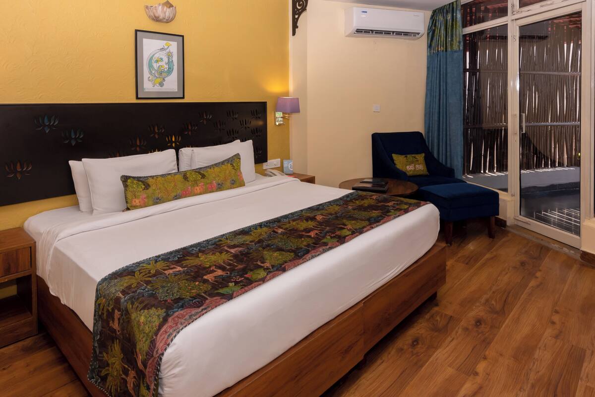 Habitación de hotel con una cama grande, cabecera de madera, mesita de noche, una silla y vistas a través de un ventanal.