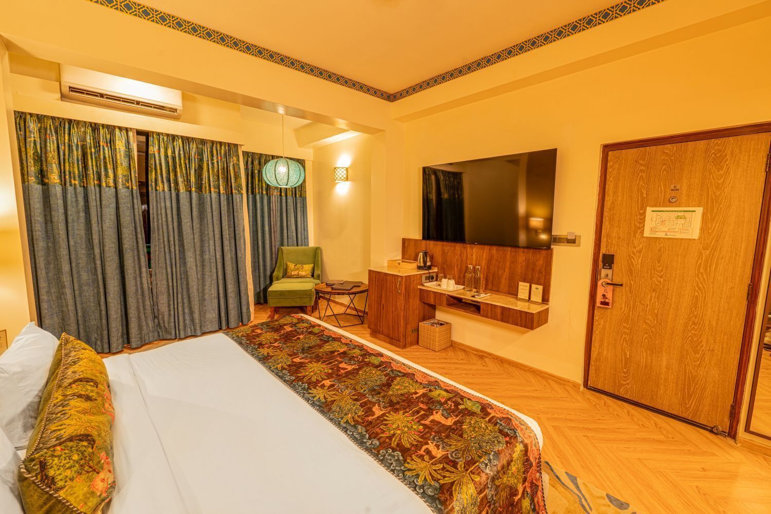A hotel room with a bed, a green armchair, a small table with a kettle, a mirror, and a wooden door.