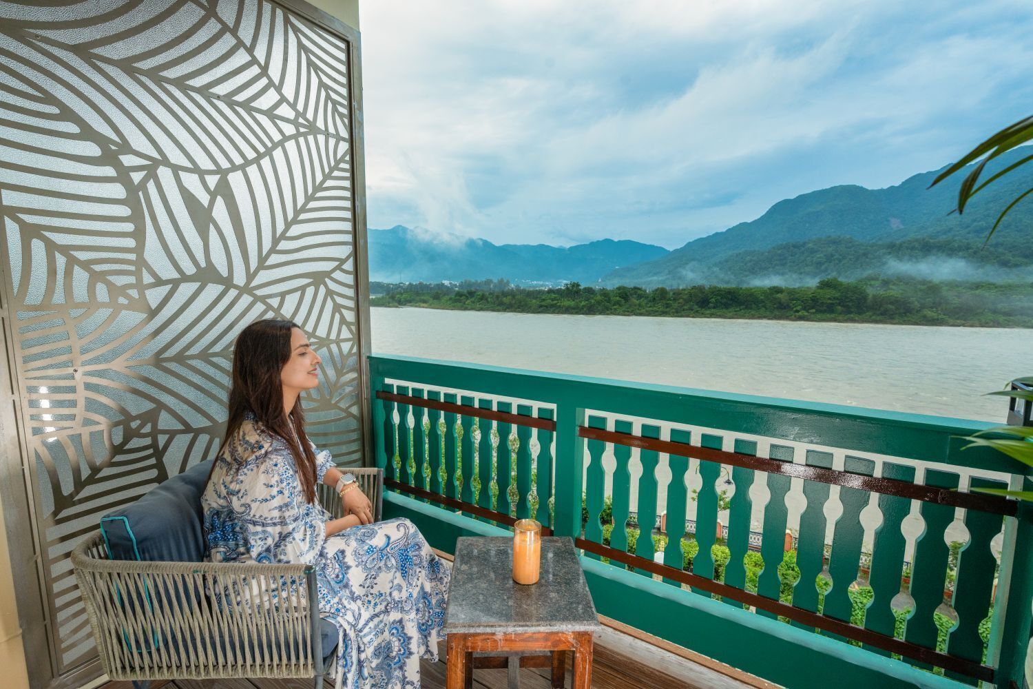 Un balcón con vista a un río y montañas, una silla de mimbre, una mesita con una vela y un panel decorativo en la pared.