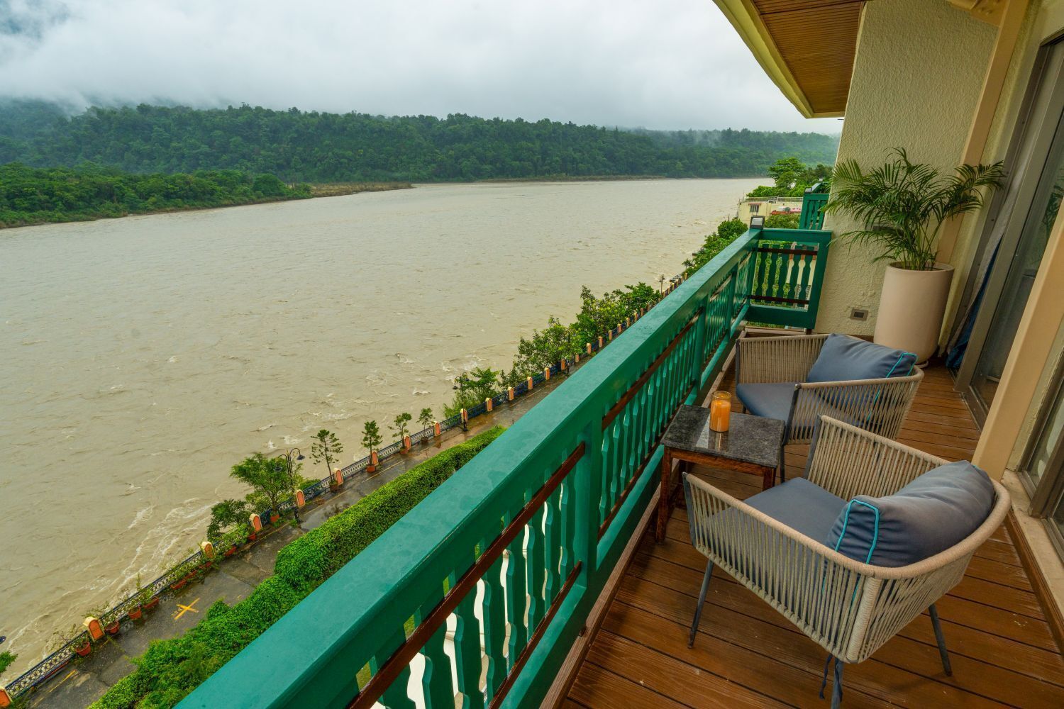 A balcony with wicker chairs and a table, overlooking a river with a forested hillside in the background.