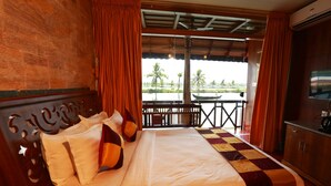 Une chambre d’hôtel comprenant un lit, une tête de lit en bois, des rideaux, un balcon avec vue sur des palmiers et un bateau, ainsi qu’un appareil de climatisation.