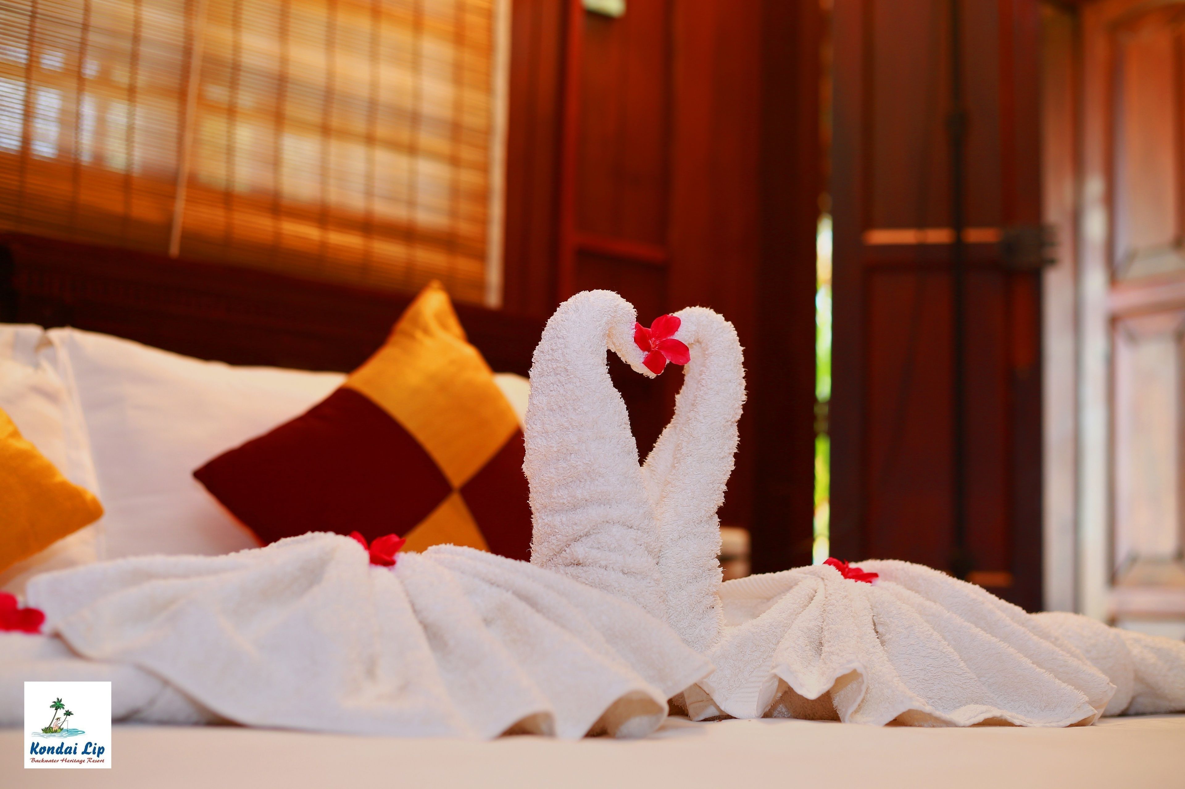 A bed with white towels arranged in a heart shape, a red flower, and a red pillow.