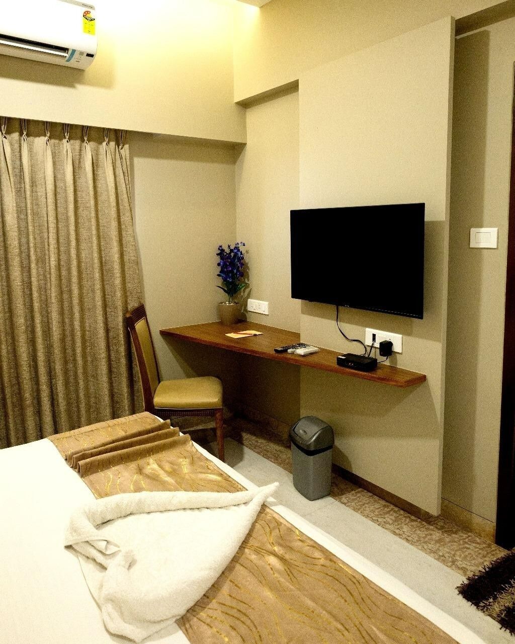 A hotel room with a bed, a desk with a television, a chair, a waste bin, and a curtain.