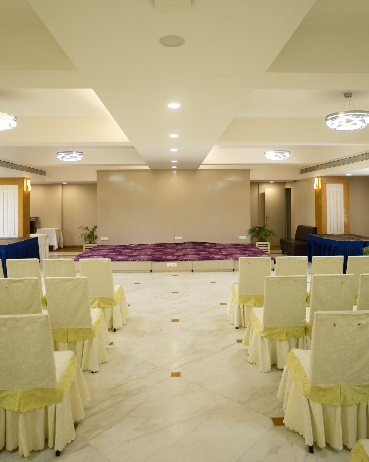 A conference room with rows of chairs, a stage with a purple carpet, and a white ceiling with recessed lighting.