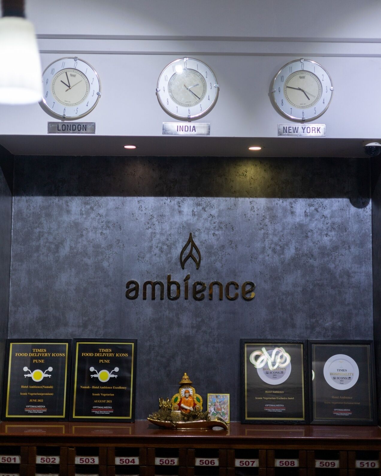 En væg med ure, der viser tiden i London, Indien og New York, samt et centralt emblem med teksten 'ambience'. Under dette er der indrammede priser for 'Times Food Delivery Icons' og 'Times Hospitality Icons'.