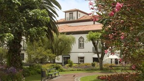 Um edifício com telhado em telhas e janelas em arco, rodeado por um jardim com bancos e plantas floridas.
