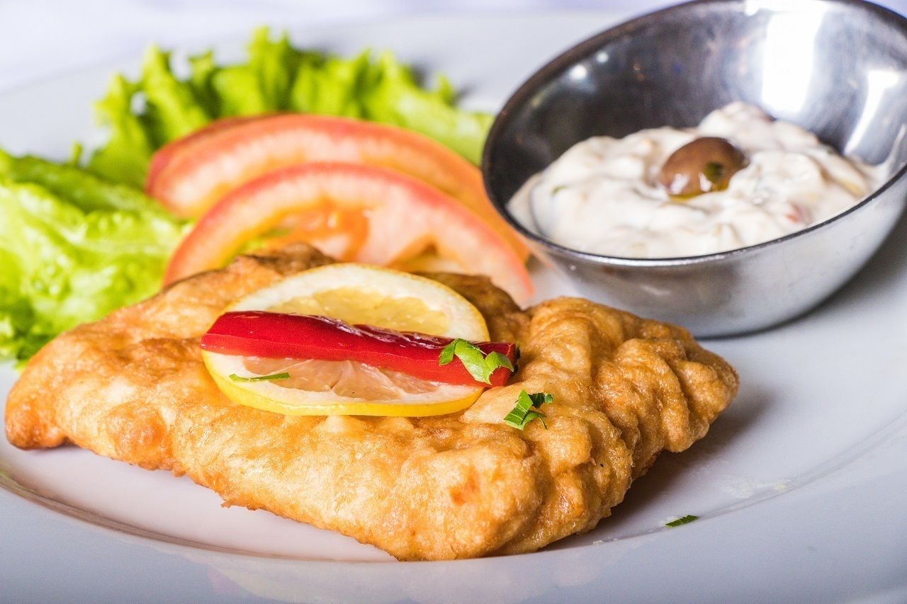 A plate with a breaded and fried item, possibly a fish fillet, garnished with lemon and red pepper, accompanied by a side of salad and a small bowl of sauce.
