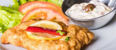 A plate with a breaded and fried item, possibly a fish fillet, garnished with lemon and red pepper, accompanied by a side of salad and a small bowl of sauce.