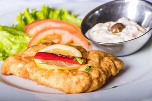 Un plato con un manjar empanizado y frito, posiblemente un filete de pescado, adornado con limón y pimiento rojo, acompañado de una ensalada y un recipiente pequeño de salsa.