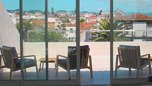 Varanda com três cadeiras de madeira e uma mesa pequena, com vista para uma paisagem urbana com edifícios de telhado vermelho e céu claro.
