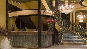 Um amplo hall com uma recepção em curva, uma escadaria com corrimão dourado e um candelabro.