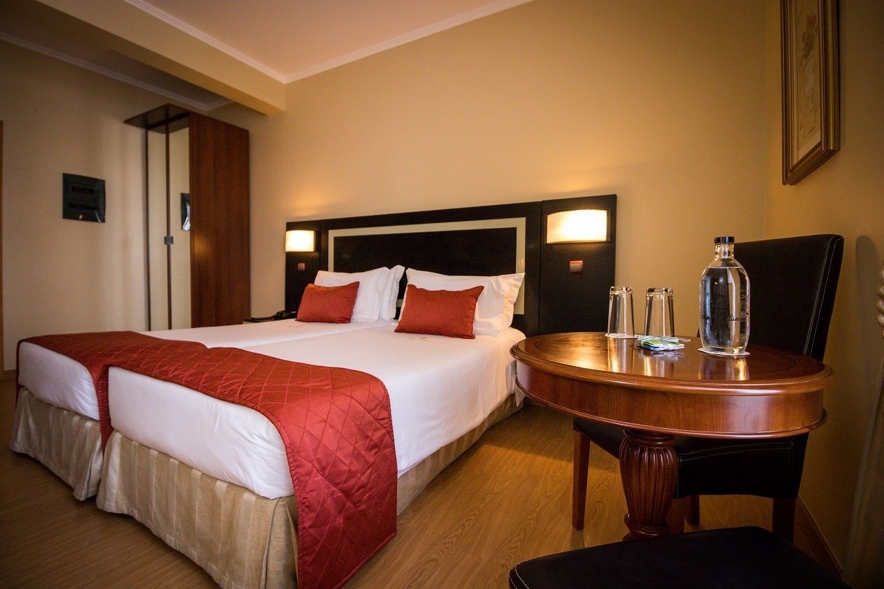 Een hotelkamer met een tweepersoonsbed, twee nachtkastjes, een houten stoel en een kleine ronde tafel met een fles en glazen.