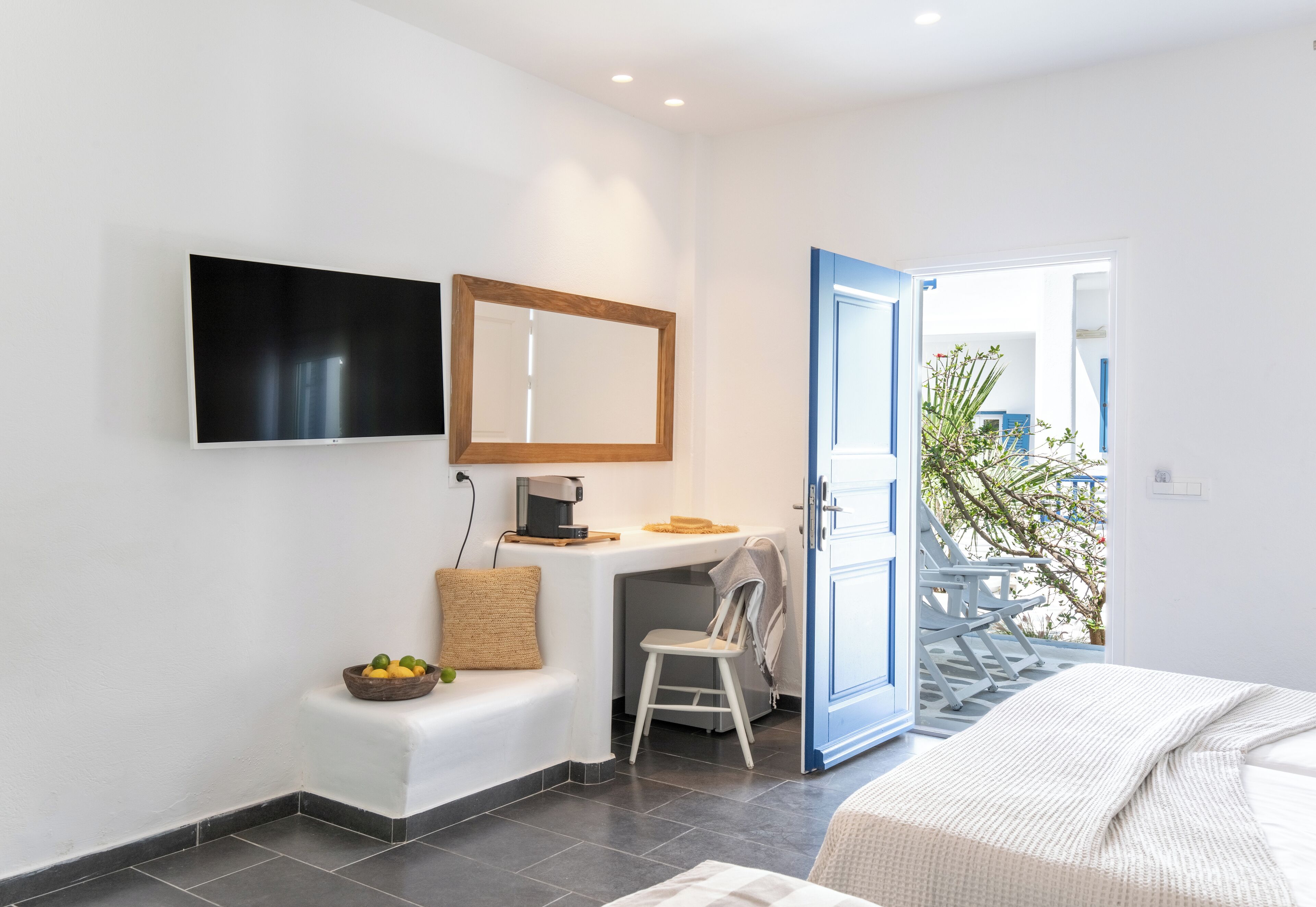 A modern hotel room with a flat-screen TV, a small desk with a coffee maker, a comfortable seating area with a cushion, and a view of the outside balcony.