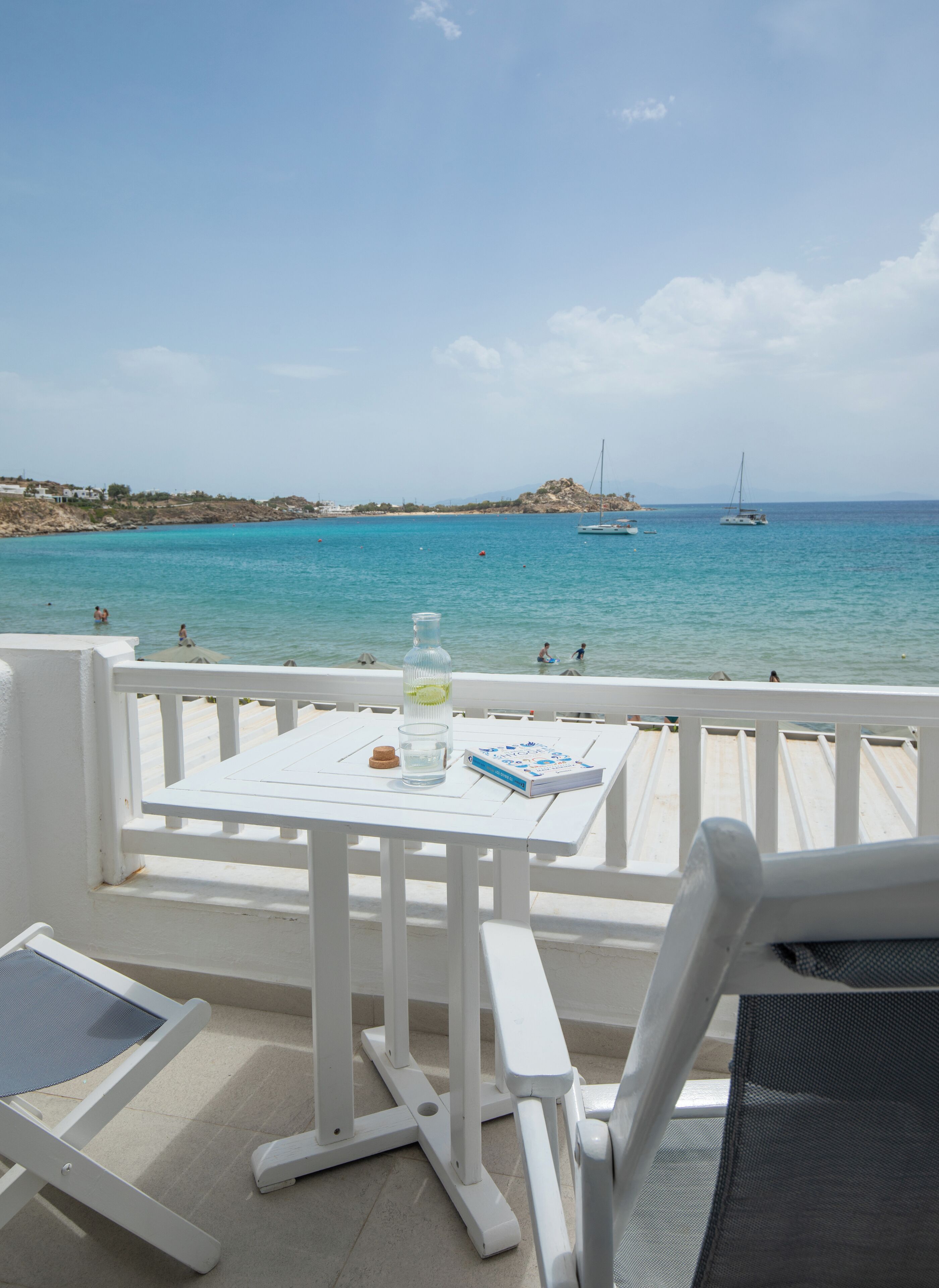 A white outdoor seating area with a table and chairs, overlooking a turquoise sea with boats and rocky coastline.