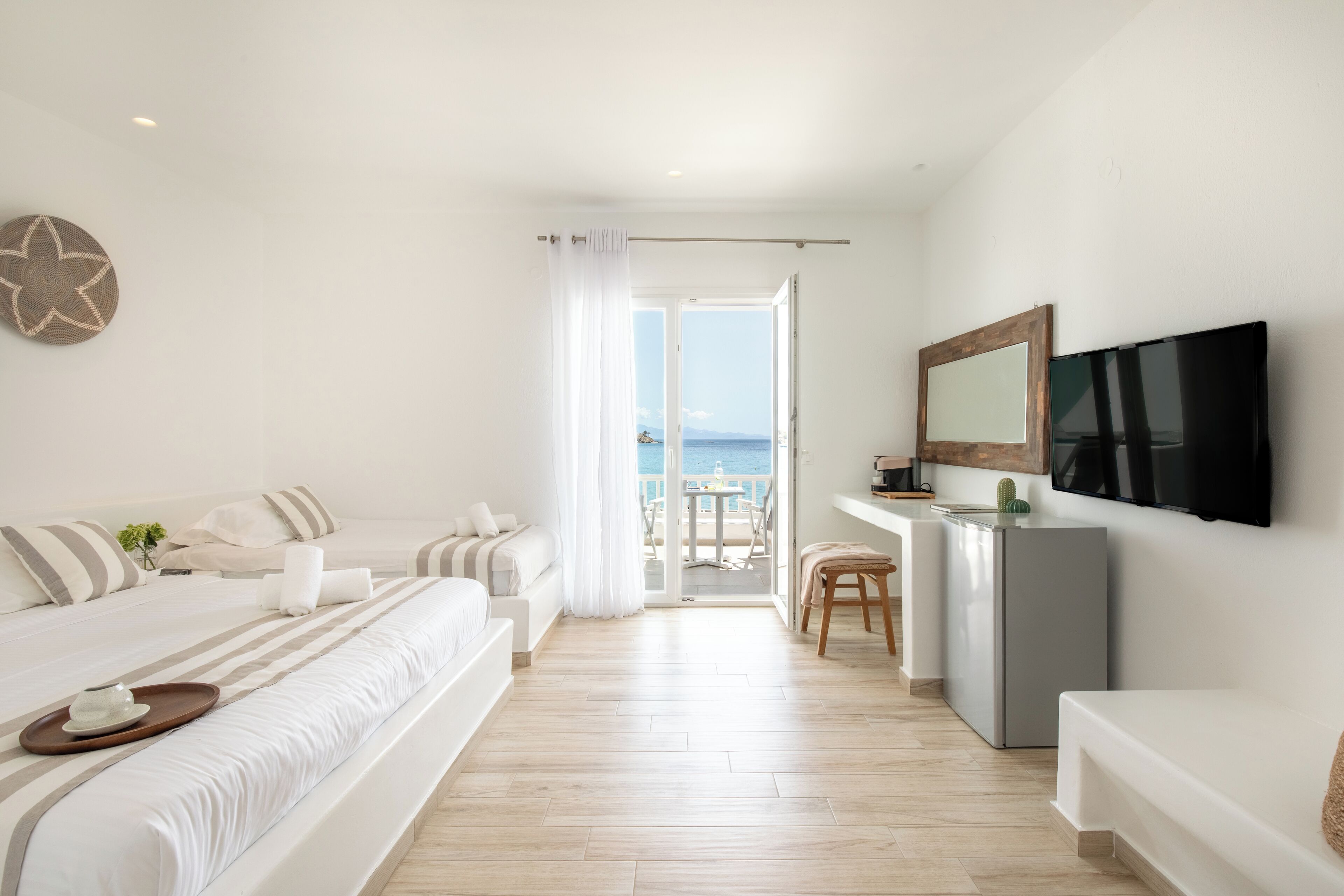 A modern hotel room with a bed, a desk, a TV, and a balcony with a view of the sea.