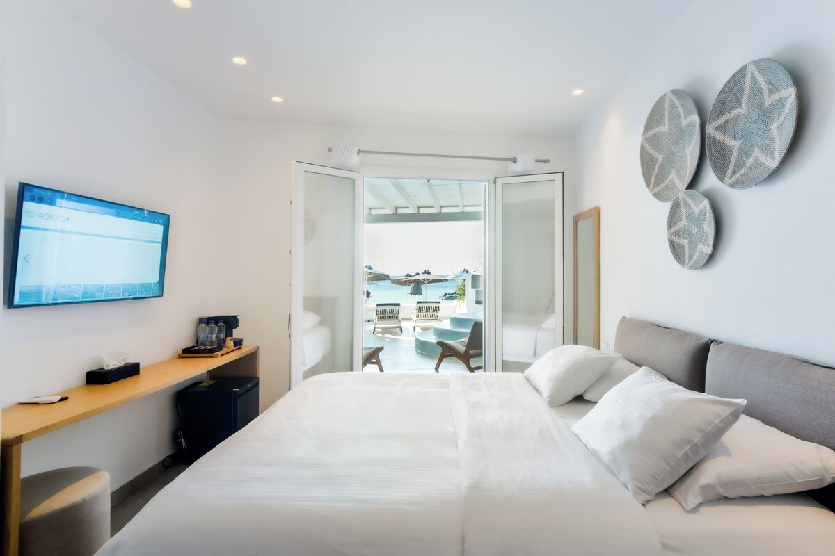 A modern hotel room with a bed, a desk, a television, and a view of the beach.