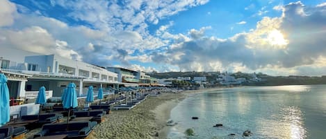 Ein Hotel direkt am Strand mit Reihen von Liegestühlen und Sonnenschirmen, einem Sandstrand und Blick auf das Meer und entfernte Gebäude.