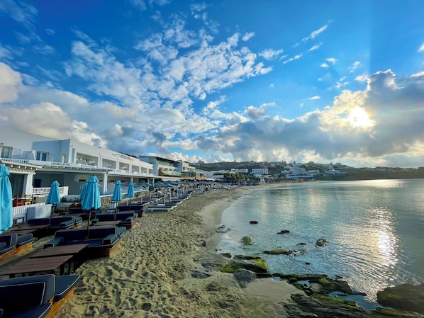 A beachside hotel with rows of sun loungers and umbrellas, a sandy beach, and a view of the sea and distant buildings. - Acrogiali Beachfront Hotel (Mykonos)