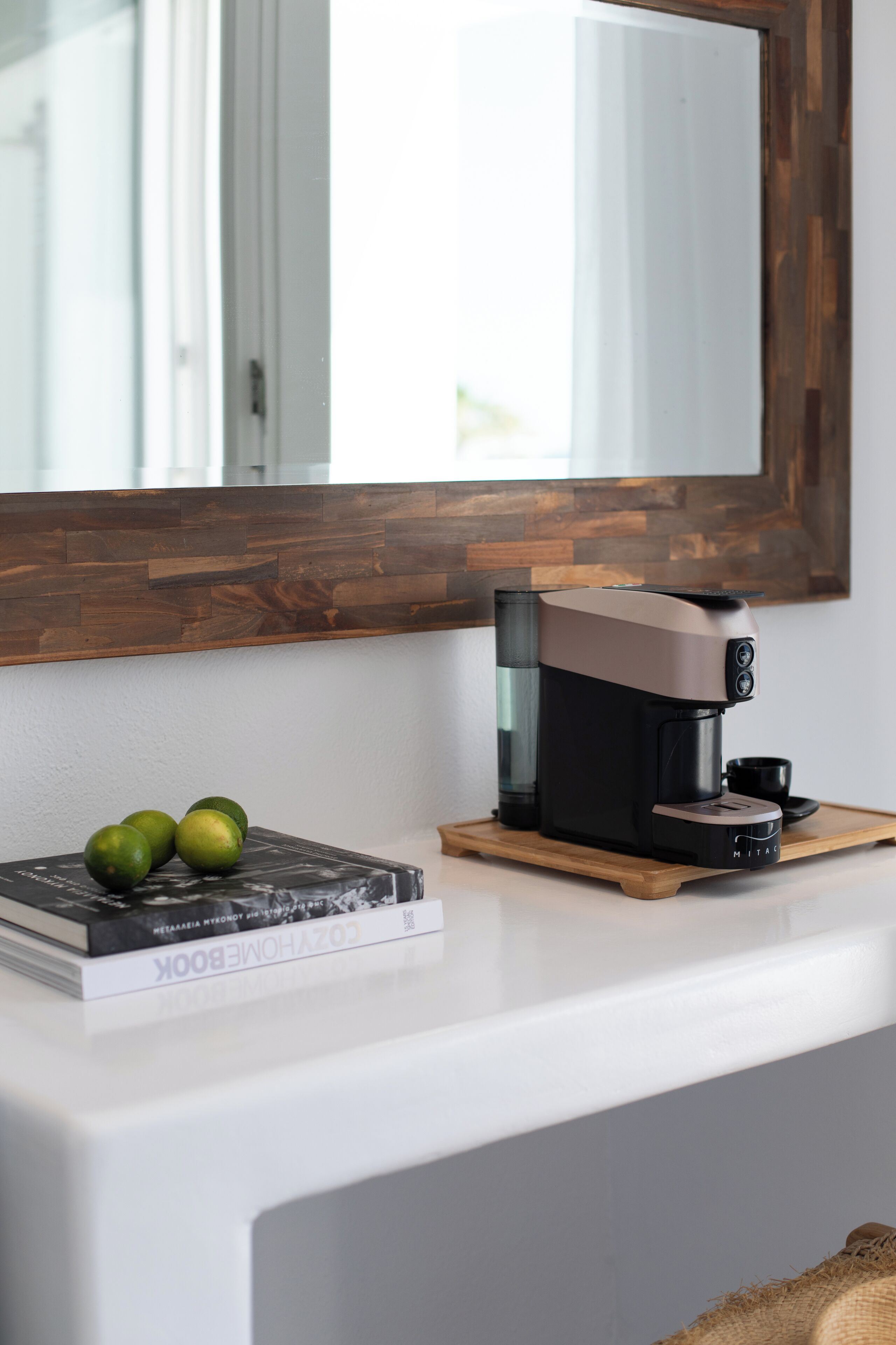 A modern coffee maker on a white countertop with a wooden tray, a book, and two limes.