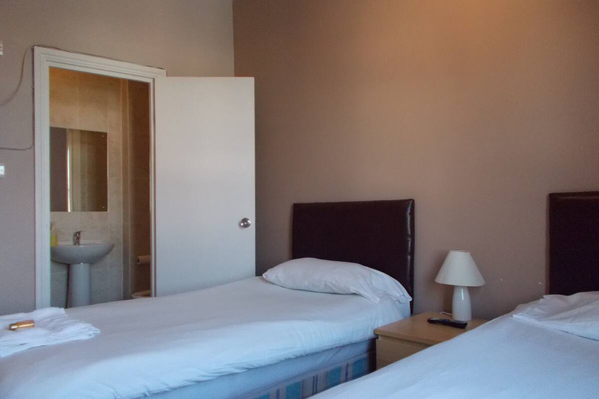 A hotel room with two beds, a bedside table with a lamp, a bathroom with a sink and mirror, and a door leading to another room.