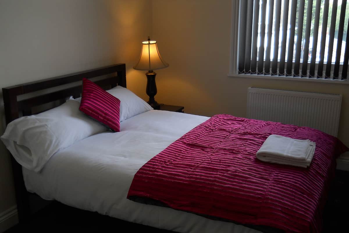 A neatly made bed with white and pink bedding, a lamp on the side, and a window with blinds.